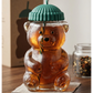 Glass bear-shaped tumbler with a green lid and straw on a wooden surface.