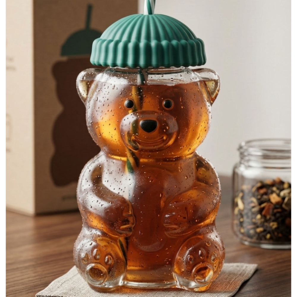 Glass bear-shaped tumbler with a green lid and straw on a wooden surface.